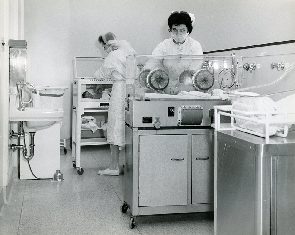 Two nurses with babies in incubators in a black and white photo.