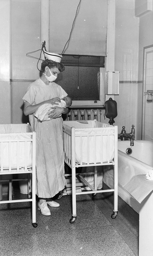 Nurse holding a baby in a small room with cribs in a black and white photo.
