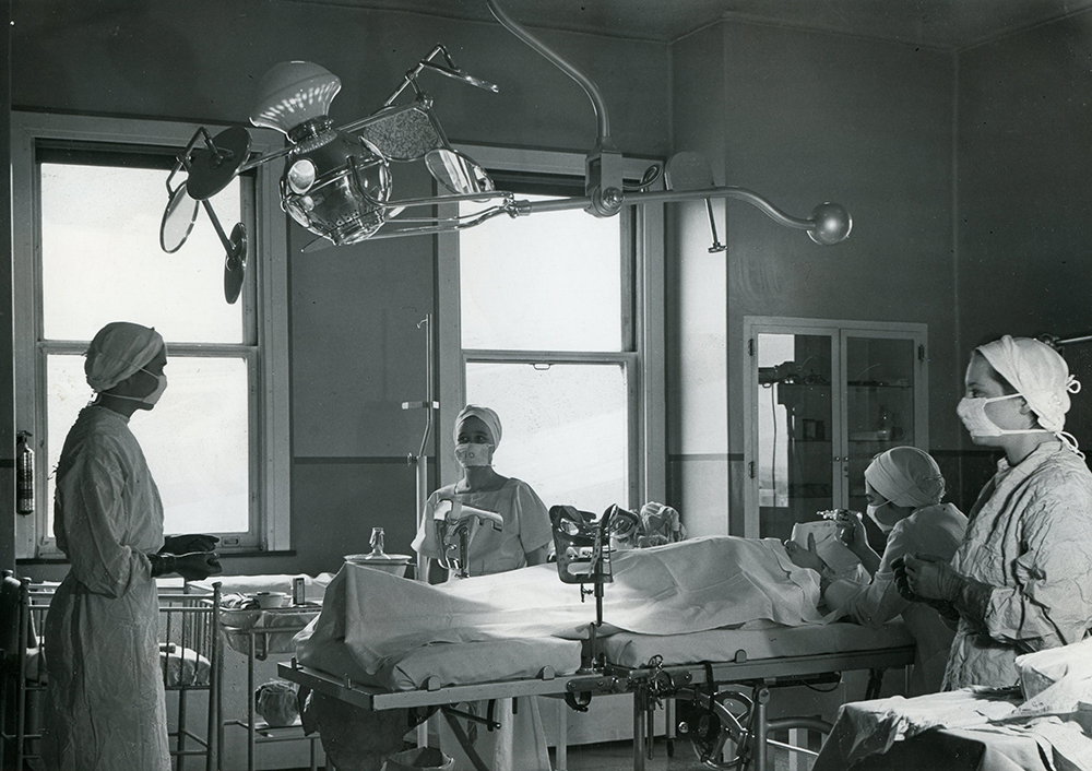 Female medical staff surround a patient on a bed in a room filled with medical equipment in a black and white photo.