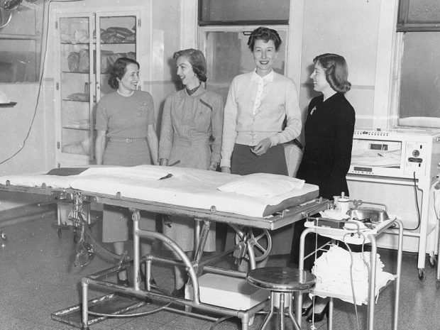 Four women in everyday clothing stand behind a gurney in a room with medical equipment in a black and white photo.