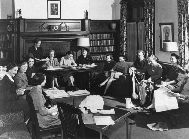 Women sit in a study engaged in paperwork and knitting in a black and white photo. Tables have been arranged by the couches for the meeting.