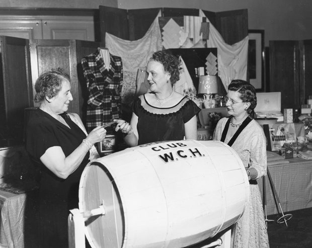 Three women draw a ballot from a barrel labelled CRADLE CLUB W.C.H. in a black and white photo. Raffle items are displayed on a table behind them.