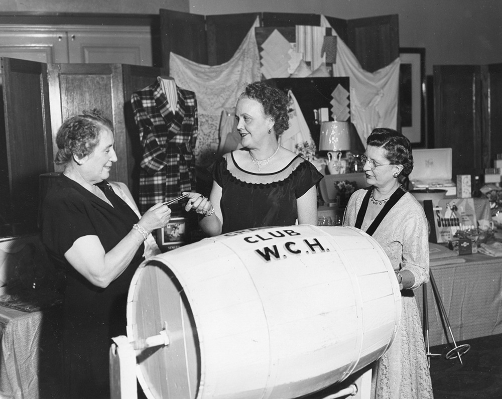 Three women draw a ballot from a barrel labelled "CRADLE CLUB W.C.H. in a black and white photo. Raffle items are displayed on a table behind them.