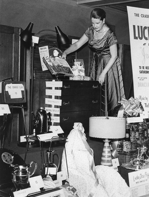 A woman in formal wear arranges domestic goods on a stage in a black and white photo.