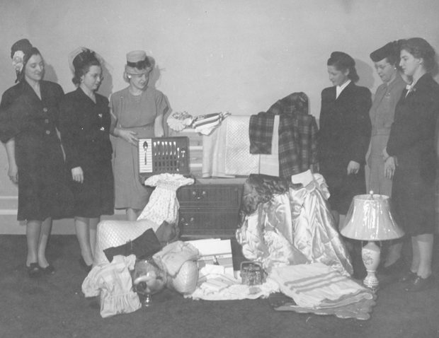 Six women in formal wear surround an arrangement of domestic goods and a large chest in a black and white photo.