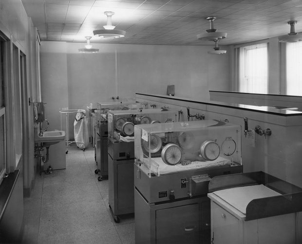 A black and white photo of a room with a row of three incubators with babies inside. A changing station is in the foreground and a sink to the left.