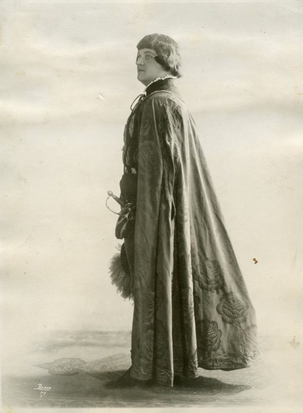 A man dressed in a costume featuring a cape and sword in a black and white photo.
