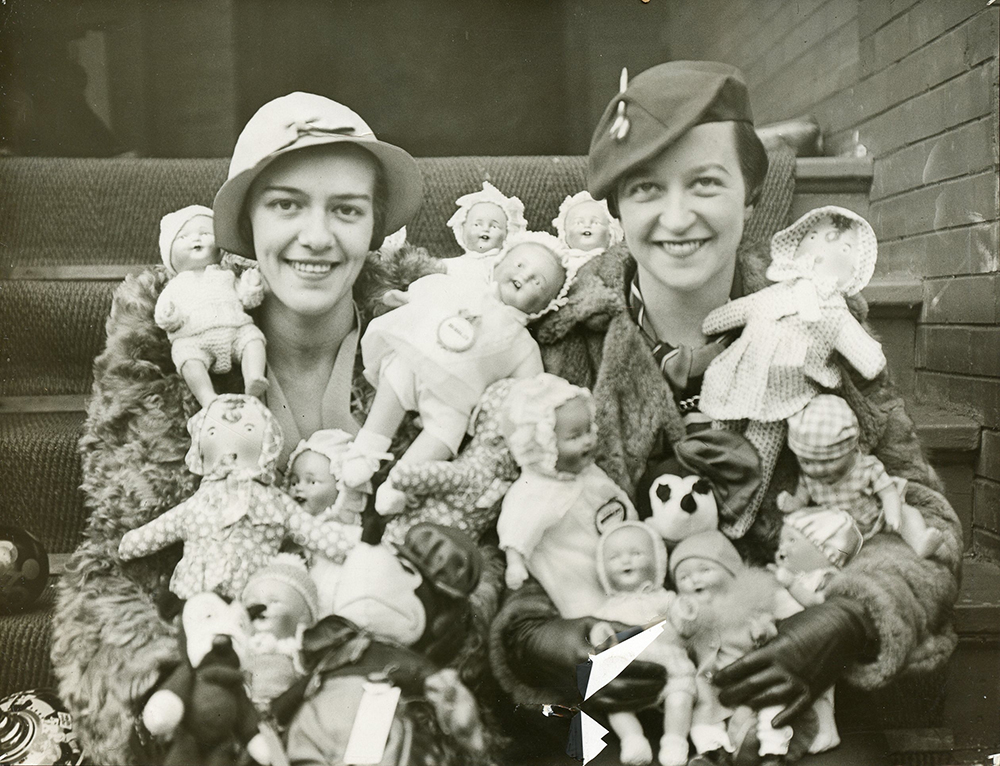 Two smiling women hold many dolls.