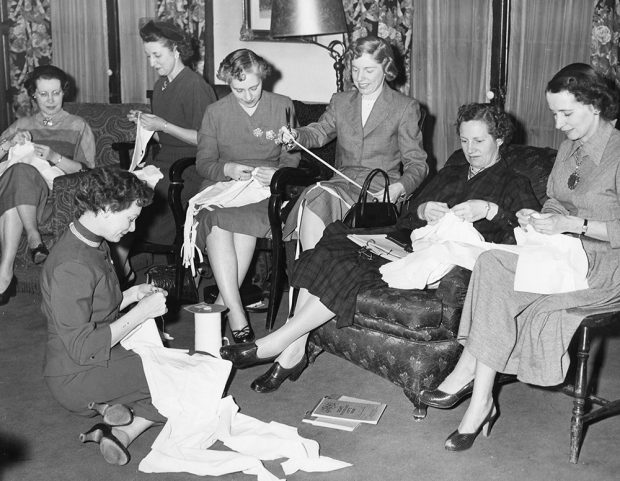 Women work on individual sewing projects in a black and white photo. Chairs have been added to the room to accommodate them. One woman sits on the floor in the left corner.