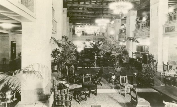 Elegantly furbished lobby with varied seating arrangements, potted palms and chandeliers in a black and white photo.