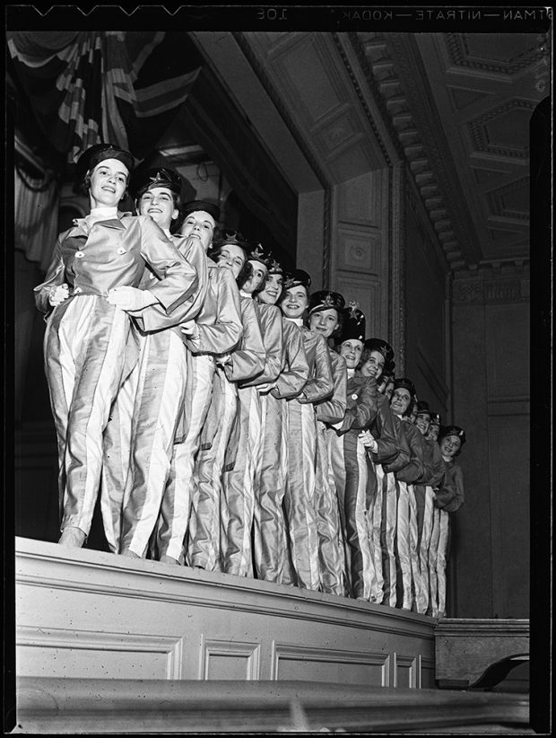 Women in matching marching band costumes pose in a row on a stage in a black and white photo.