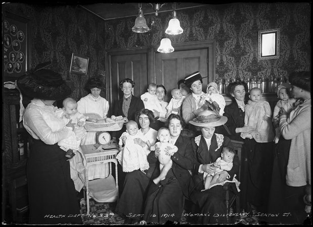 A large group of mothers holding their babies are posed in a parlour in a black and white photo. To the left is a nurse weighing a baby.