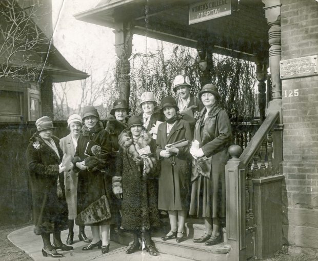 A group of fashionably dressed women stand outside a building with a small sign reading Women's College Hospital in a black and white photo.
