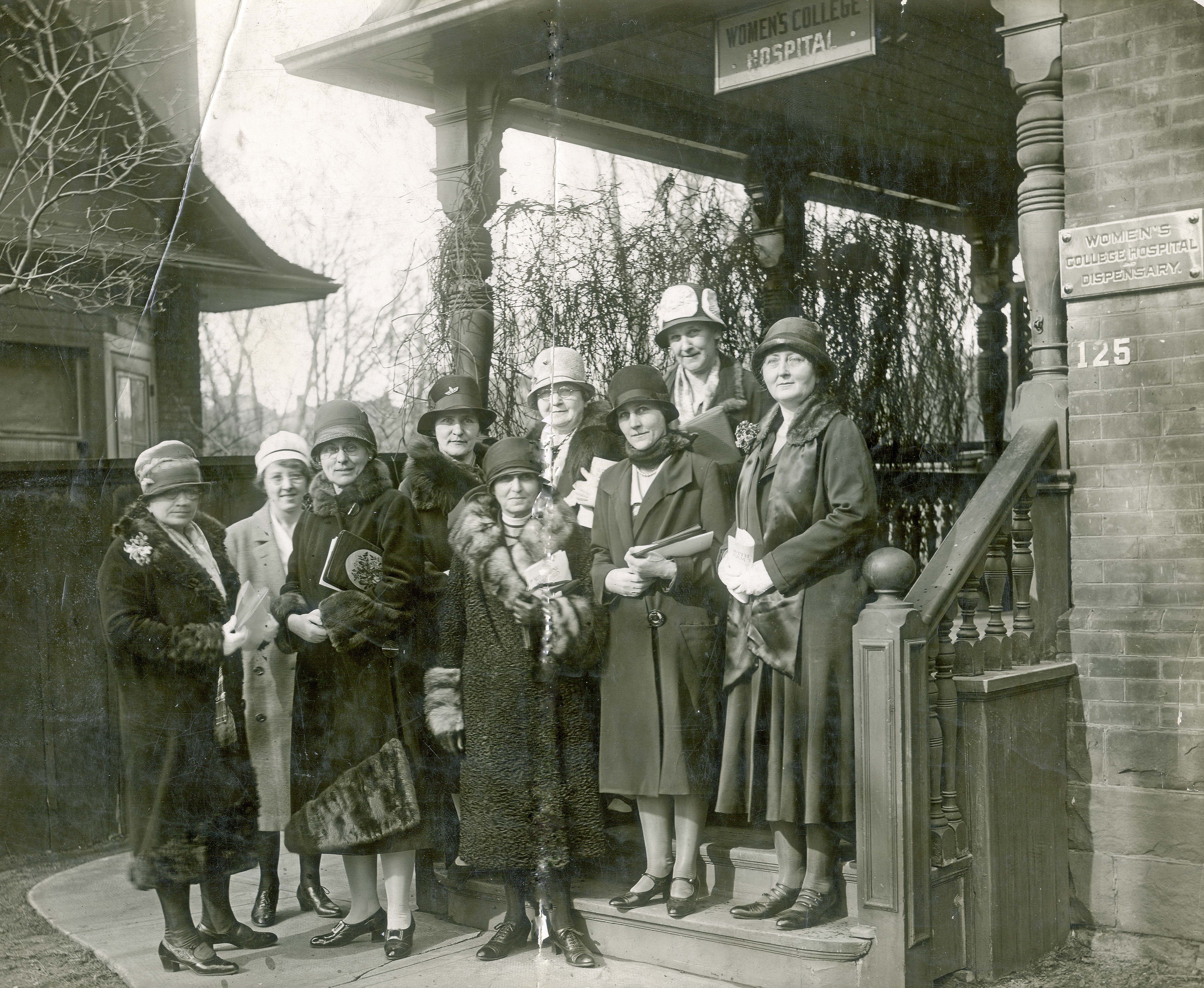 A group of fashionably dressed women stand outside a building with a small sign reading "Women's College Hospital" in a black and white photo.
