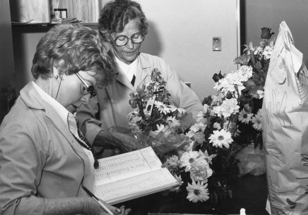 Two women; one notes in a ledger, the other arranges a bouquet in a black and white photo.