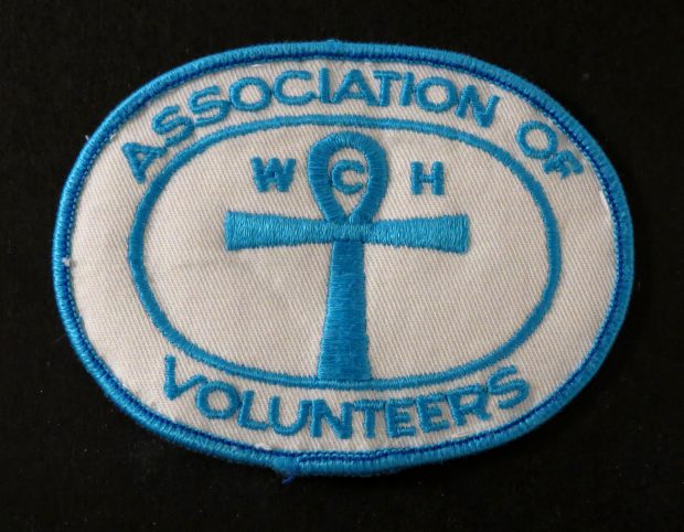 White badge with blue stitching. Text reads: Association of Volunteers.