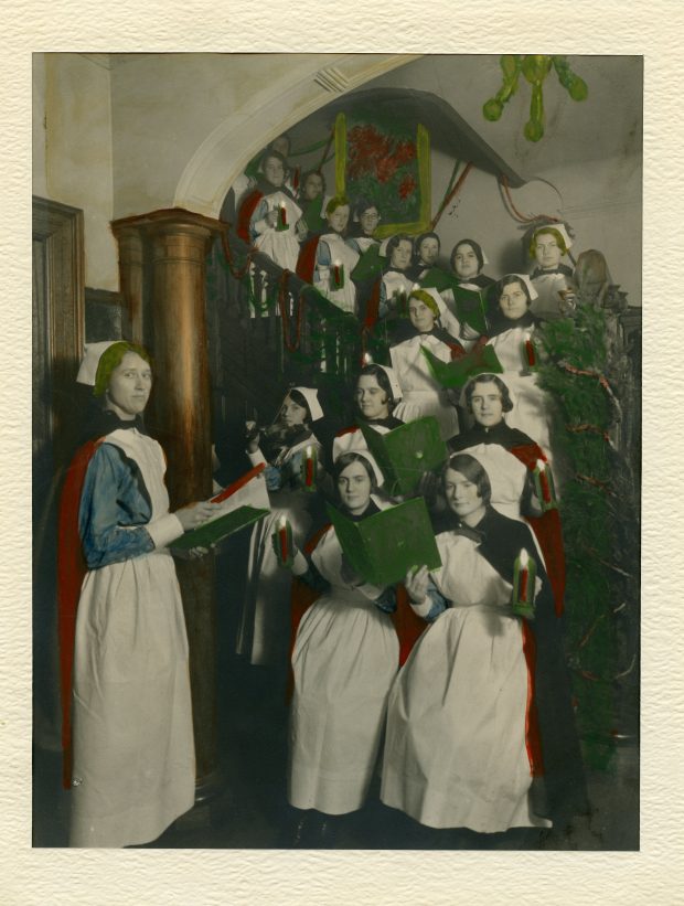 Nurses holding candles and carol books are lined up on the stairs in an old photo. One nurse leads them while another plays violin.