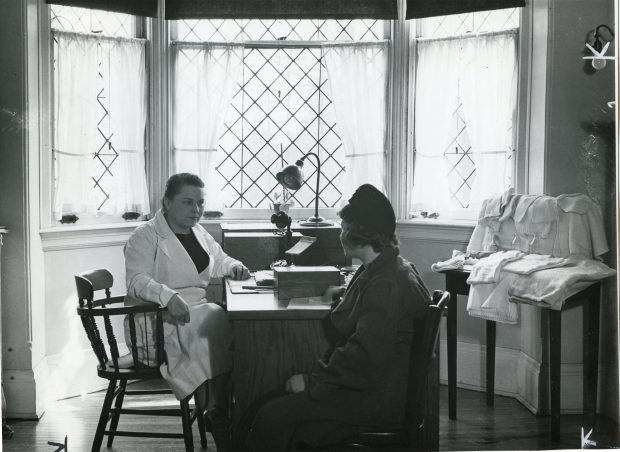 A medical professional consults with a woman at a desk in front of a bay window in a black and white photo. Baby linens and knitted clothes are displayed on a side table on the right.