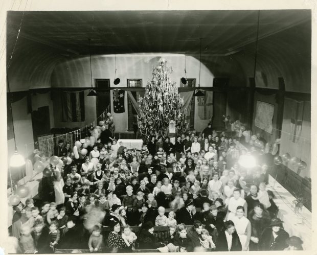 Women, children and nurses gather around long tables in a black and white photo. Balloons are hung and Santa stands before a large Christmas tree at the back of the room.