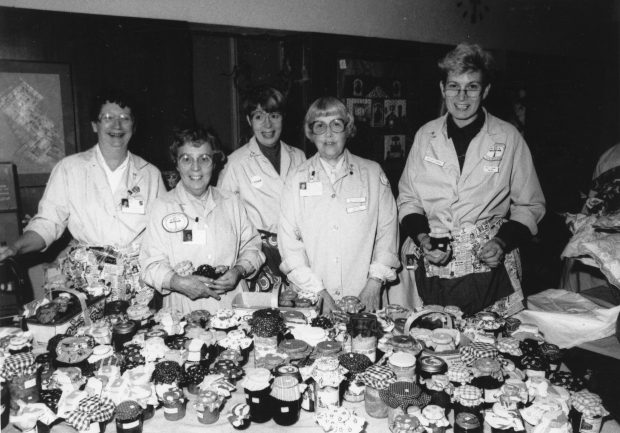Women in matching overcoats stand before a table laden with preserves in a black and white photo.