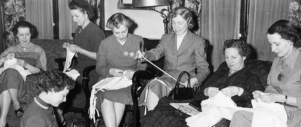 Women work on individual sewing projects in a black and white photo. Chairs have been added to the room to accommodate them. One woman sits on the floor in the left corner.