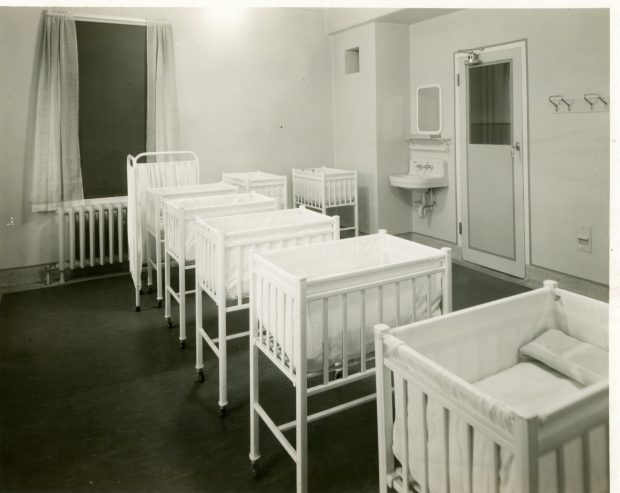 Multiple cribs in a bright, open room in a hospital with a sink and mirror in the corner next to the door, in a black and white photo.
