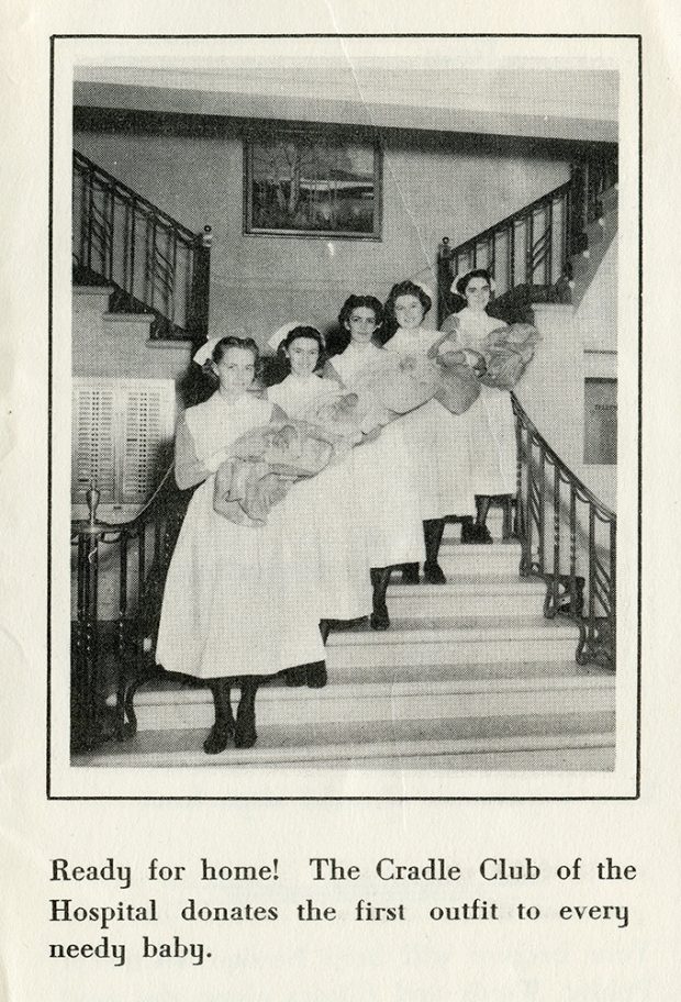 Five young nurses pose on a staircase proudly holding babies wearing hats and wrapped in blankets in a black and white photo.