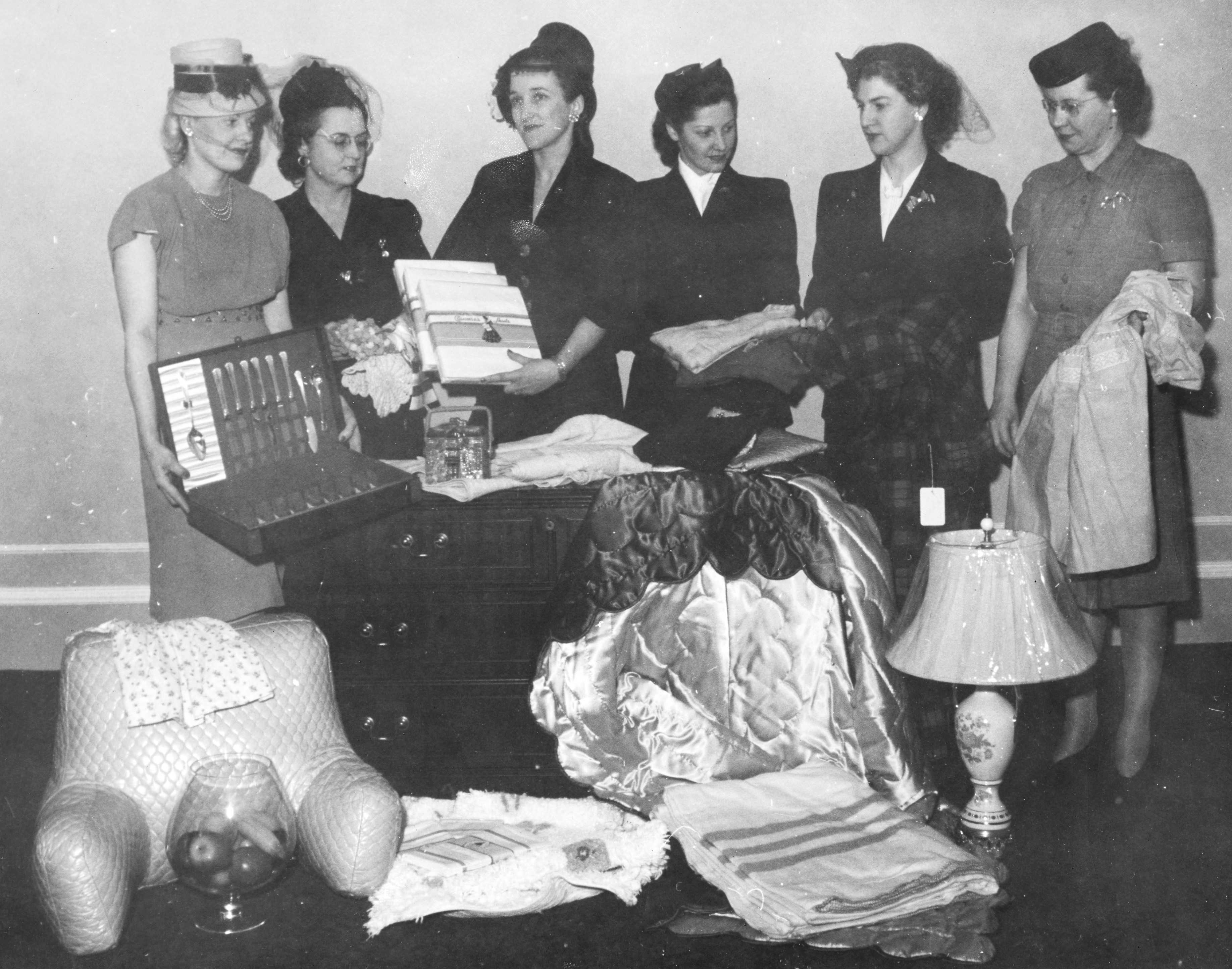 Six women in formal wear stand behind an arrangement of domestic goods in a black and white photo.