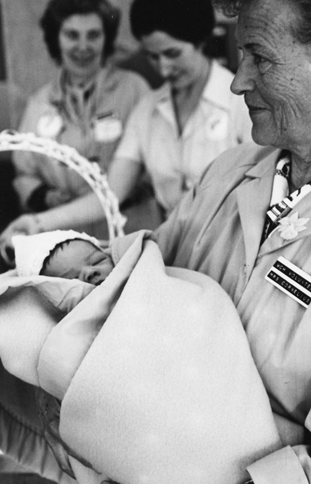 In a black and white photo, a woman in volunteer coat holds sleeping baby wrapped in a blanket; two more women in the background.
