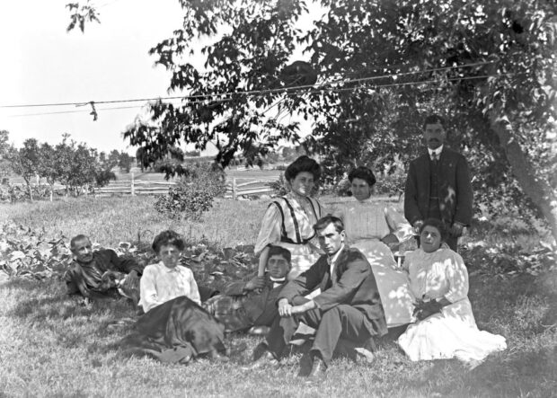 Old photo of a field bordered by apple trees and eight people seated on the ground