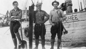 Photographie en noir et blanc. Trois hommes se tiennent devant la goélette Josuhée. Celui de gauche tient une pelle, celui du centre une longue scie et celui de droite, une hache. 