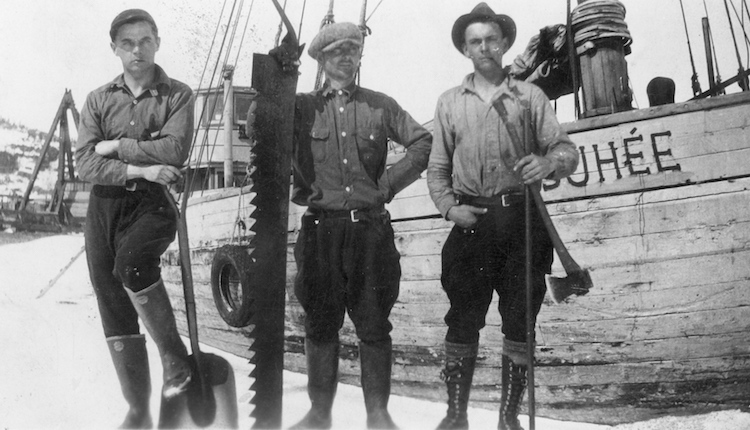 Black and white photograph Three men standing in front of the Josuhée schooner. The person on the left is holding a shovel, the one in the centre is holding a saw and the one on the right is holding an axe.