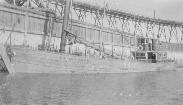 An old black and white photograph of the Josuhée. The wooden ship has its sail lowered, with its name clearly visible. At the back, Captain Joseph Desgagnés is standing proudly outside his cabin. There is a wooden dock in the background.