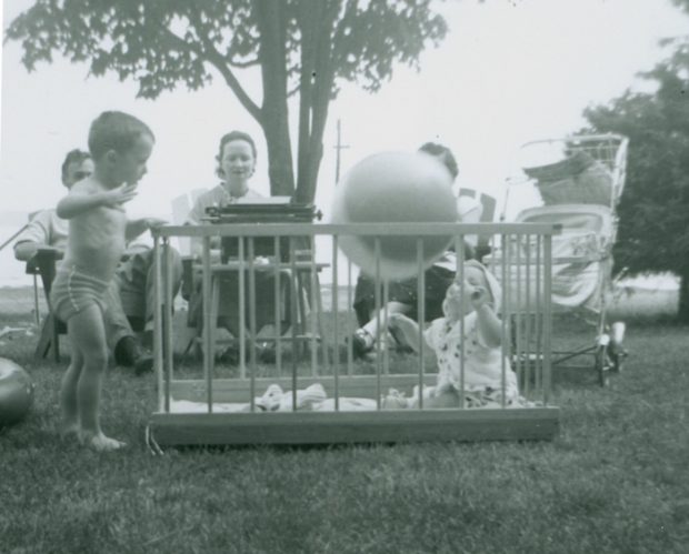 This black and white photograph, taken outdoors, shows a baby in a park playing with a ball in the foreground. Left of the child, a young boy is standing on the grass. Three adults are sitting in the back. The woman in the middle is typing on a typewriter resting on a small table in the grass.