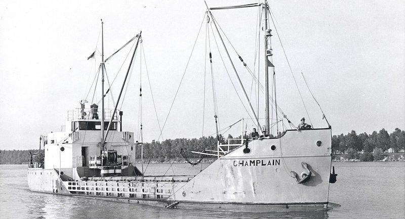 Photographie en noir et blanc. On observe le caboteur Champlain dans toute sa longueur, vue de devant. Le bateau est pâle et son nom est inscrit en lettre noire à la proue. Il a deux mâts de charge et ne semble transporter aucune cargaison. Trois hommes se tiennent au-devant du bateau qui flotte sur l’eau.