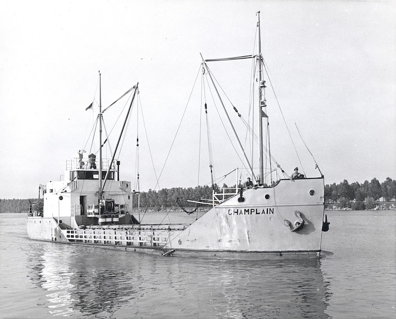 Black and white photograph. This picture gives us a front view of the Champlain coaster in all its length. The ship is light-coloured, with its name written in black lettering on its prow. It is also fitted with two boom masts but does not seem to be carrying any cargo. Three men are standing at the front of the ship, which is floating on the water.