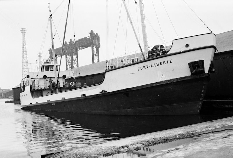 Black and white photograph. This picture gives us a front view of the Fort Liberté coaster in all its length. The ship’s hull is painted black and white, with its name written in black lettering on its prow. It is also fitted with two boom masts but does not seem to be carrying any cargo. Part of a lock canal is visible in the background.