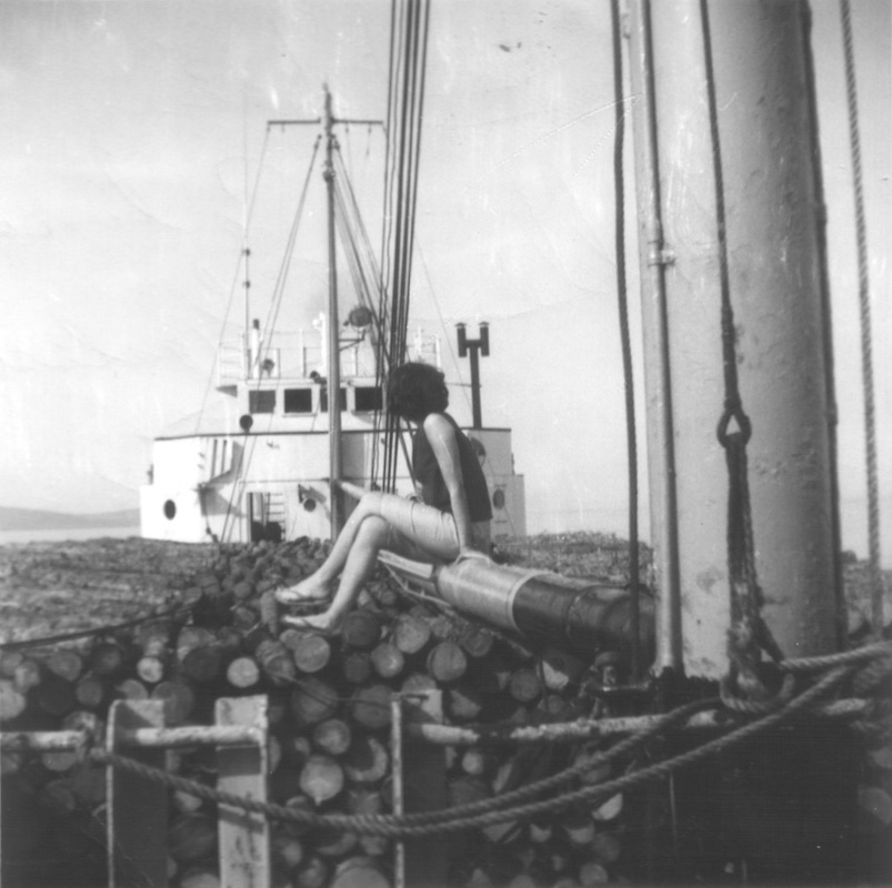 A black and white photograph taken on the deck of the ship, at stern. The ship is filled with rigging, on top of which a woman is seated, facing away. In front of her, the boom mast and white ship’s cabin are visible.