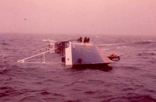 Colour photograph of a half-submerged l’Aigle d’Océan. The photograph was taken in front of the ship’s hull. The ship is lying on its side with its masts immersed in water. Crew members are waiting to be rescued on the blue and yellow hull of the ship.