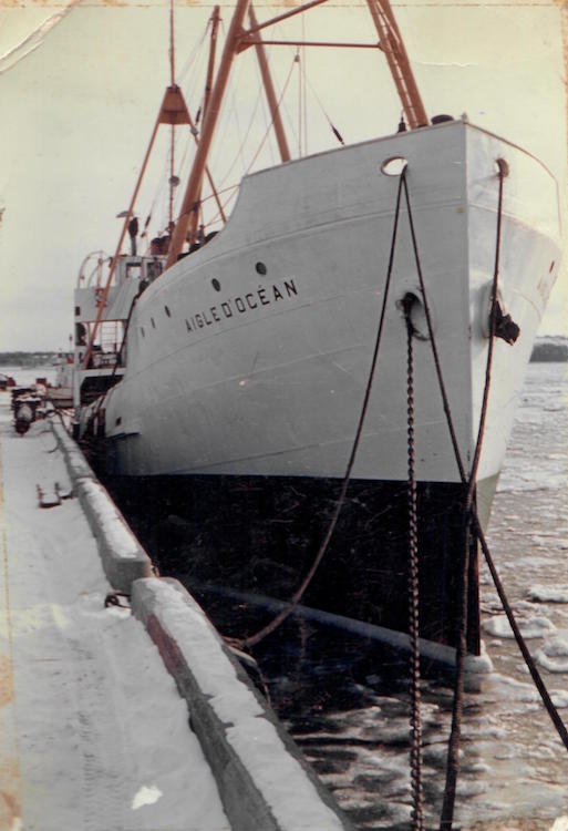 A photograph of the ship l’Aigle d’Océan at dock, front view. The ship is white but has a black stripe on the lower portion of its hull. The name of the ship appears on the upper portion of the hull, just under the portholes. There are little clumps of ice in the water.