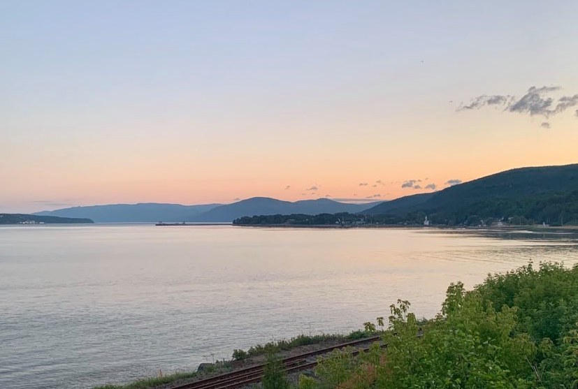 View of the St. Lawrence River at sunset. In the foreground, a section of train track and some bushes. In the background, a body of water with pink reflections. A few clouds speckle a pink and blue sky. There are mountains visible in the distance.