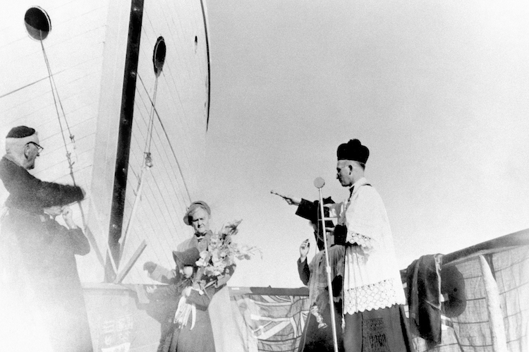 This black and white photograph shows six people: an elderly woman and a child holding a bouquet of flowers (centre) with priests and altar boys around them. A ship’s hull appears in the background.