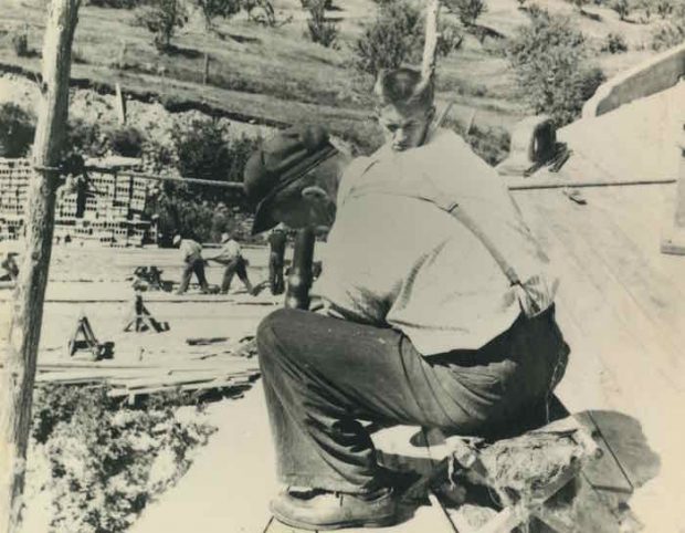 Black and white photograph. Two people are in the foreground. They appear to be sitting on the hull of a ship. One has a mallet in his hand. Just behind him, a young man watches him carefully. In the background one can make out a construction site, complete with various construction materials and workers.