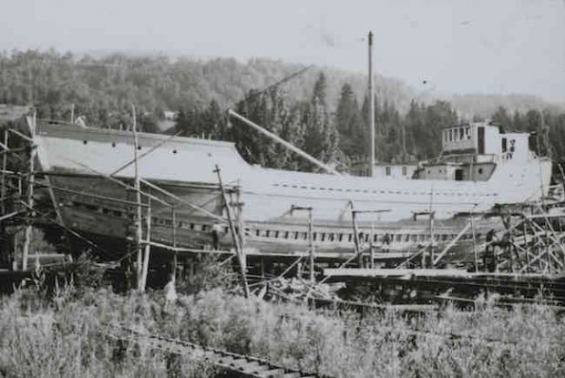 This black and white photograph shows a schooner under construction. The ship appears to be almost finished. There is wooden scaffolding all around the structure. A few men—tiny in the photograph—are busy working on the ship. In front of the ship, a man is standing in the tall grass observing the scene.