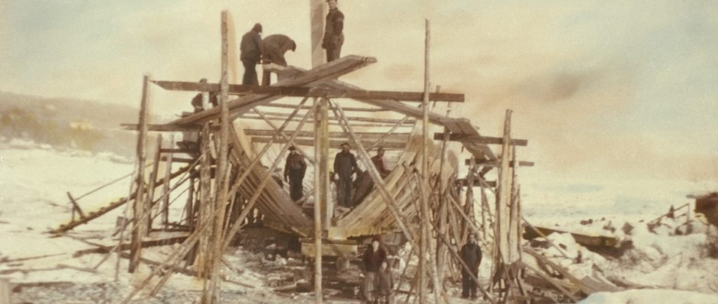 View of a schooner under construction. The structure of the ship is surrounded by scaffolding. There are men in work clothes standing on the hold of the ship and the scaffolding. A woman, a child and another man are standing in the foreground. This photograph was taken in winter. The photo has been coloured to give it a sepia tone.