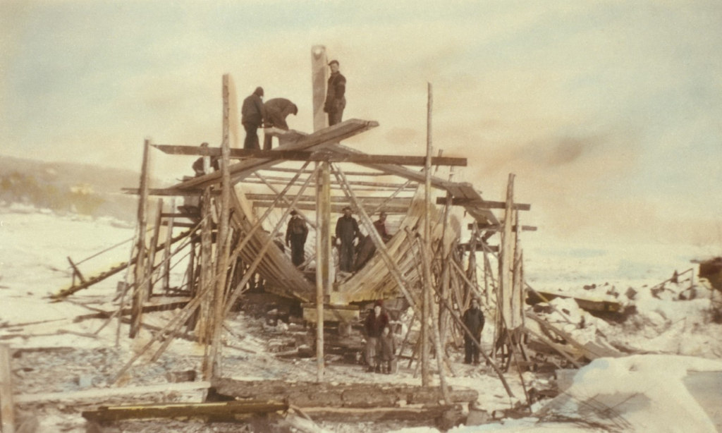 View of a schooner under construction. The structure of the ship is surrounded by scaffolding. There are men in work clothes standing on the hold of the ship and the scaffolding. A woman, a child and another man are standing in the foreground. This photograph was taken in winter. The photo has been coloured to give it a sepia tone.
