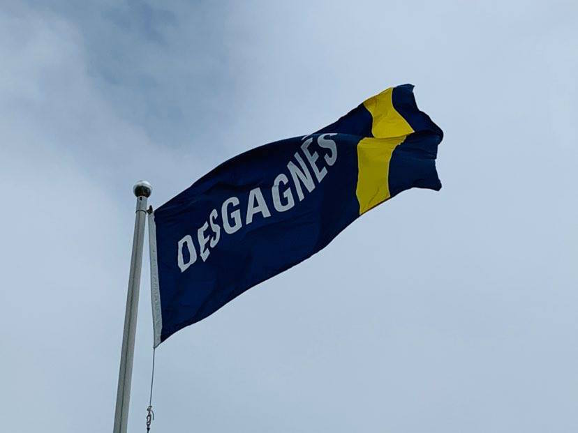 The flag of Groupe Desgagnés floating in the breeze. The name Desgagnés is written in white on a navy-blue background. There is a yellow vertical stripe across the right side of the flag.