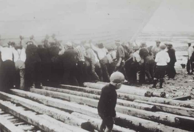 An old black and white photograph. In the foreground, a young child walks alone on wooden planks. In the background, around two dozen men are pushing a schooner towards the river.