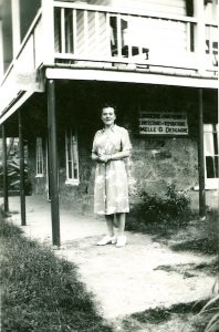 Black and white photograph. A woman wearing a light-coloured dress with a motif is standing under the deck of a house. The lower portion of the house has stone walls, whereas the upper portion has wooden walls. A sign is affixed on the stone wall reading “Melle G.Desgagne” (Miss G.Desgagne).