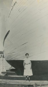 A black and white photograph. A woman wearing a light-coloured dress is standing in front of a wooden ship. The woman looks tiny next to the massive ship. In the upper left corner of the photograph, we can see the name of the ship painted on the hull: Mont Royal.